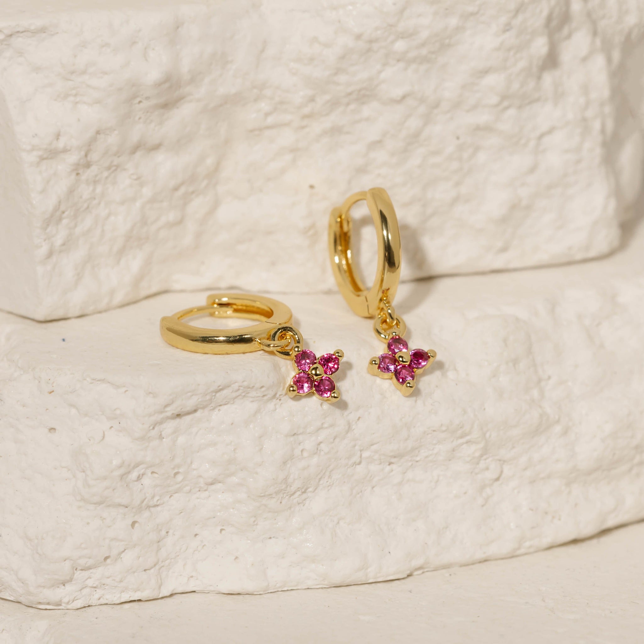 A pair off Clover Drop Hoop-Fuchsia earrings is displayed atop two tiered stone slabs, the crystal charms and golden hoops gracefully reflecting the light.