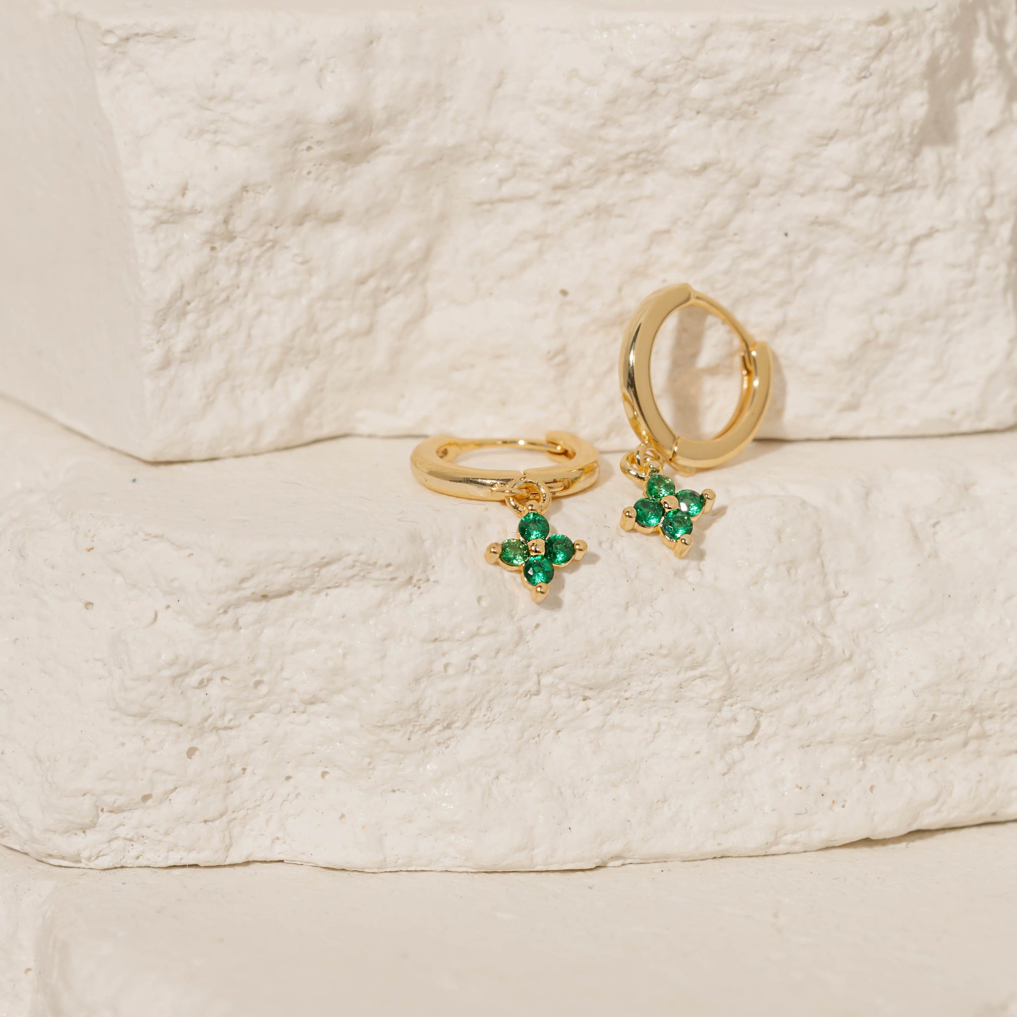 A pair of Clover Drop Hoop-Verde earrings sits against tiered stone slabs, the golden hoops and green crystals sparkling in the light. 