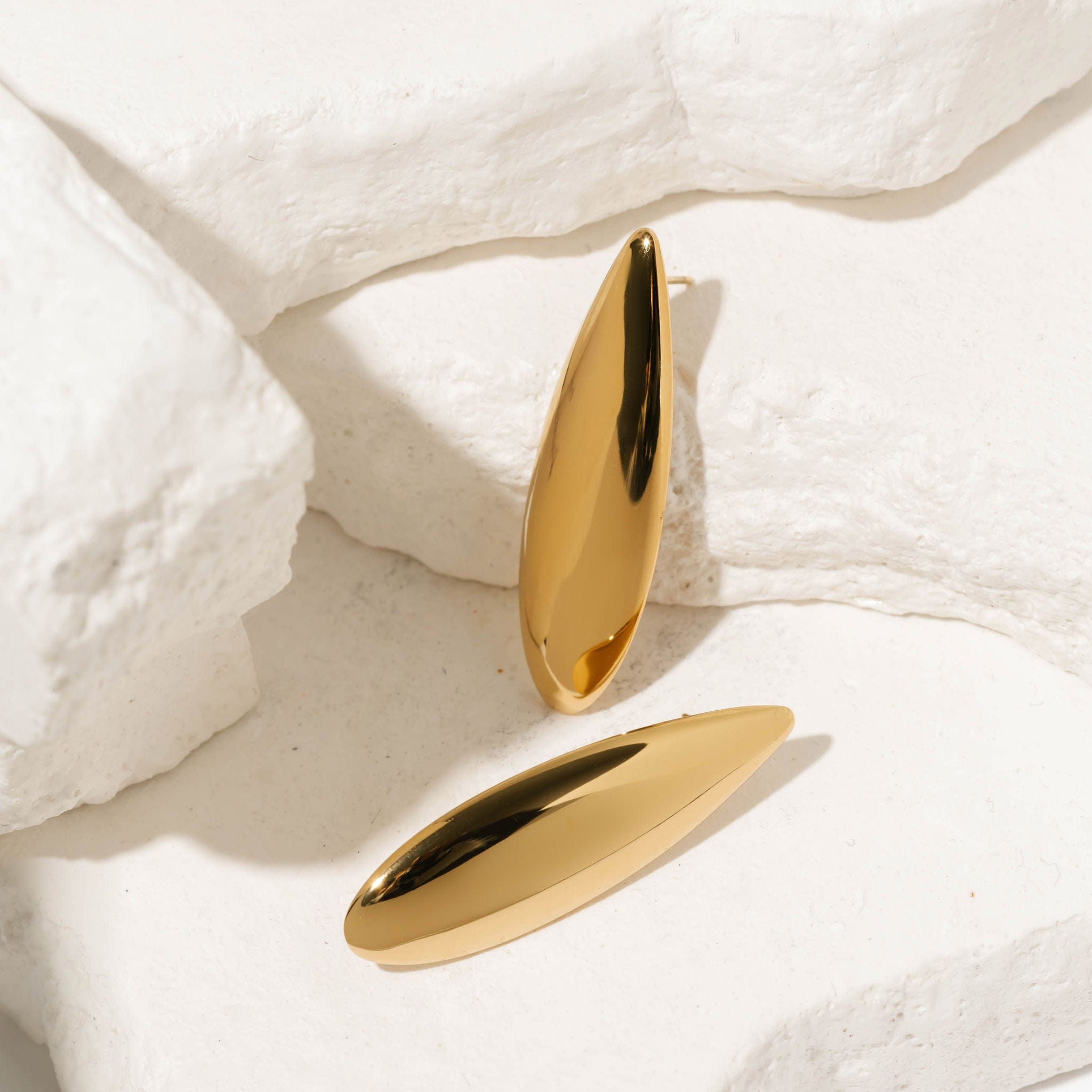 The Perfect Hoop Lotus Earrings in gold displayed on white textured stone backdrop, showcasing their sleek curved teardrop design in a flat lay.