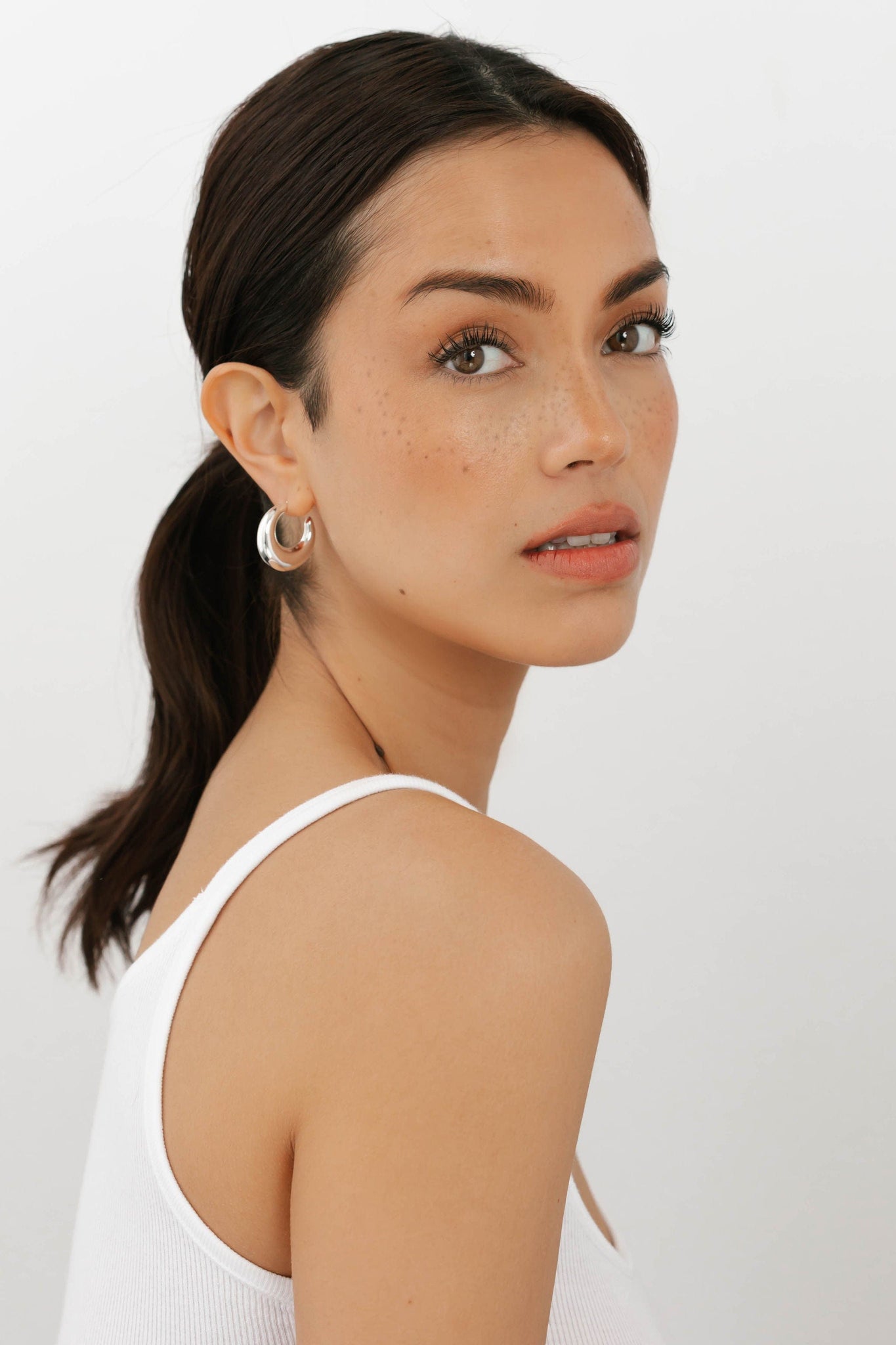 Model wearing a white tank top and silver crescent hoop earrings by The Perfect Hoop, against a plain background.