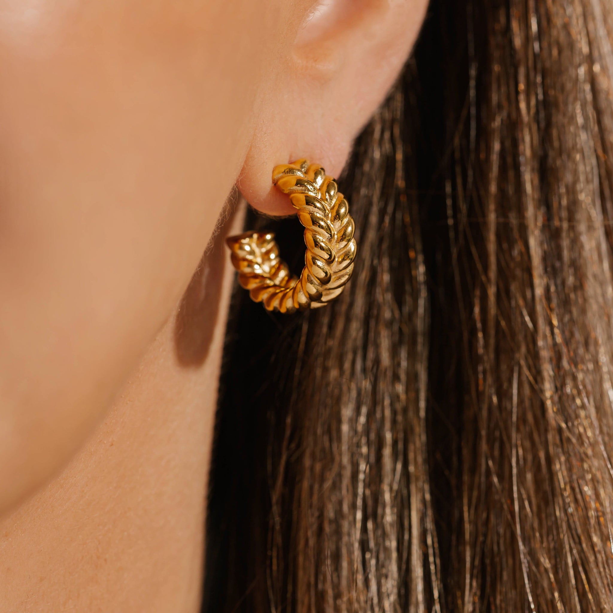 The Trenza Hoop's broad, golden hoop with its braided open hoop design looks stunning as it decorates the model's ear. 