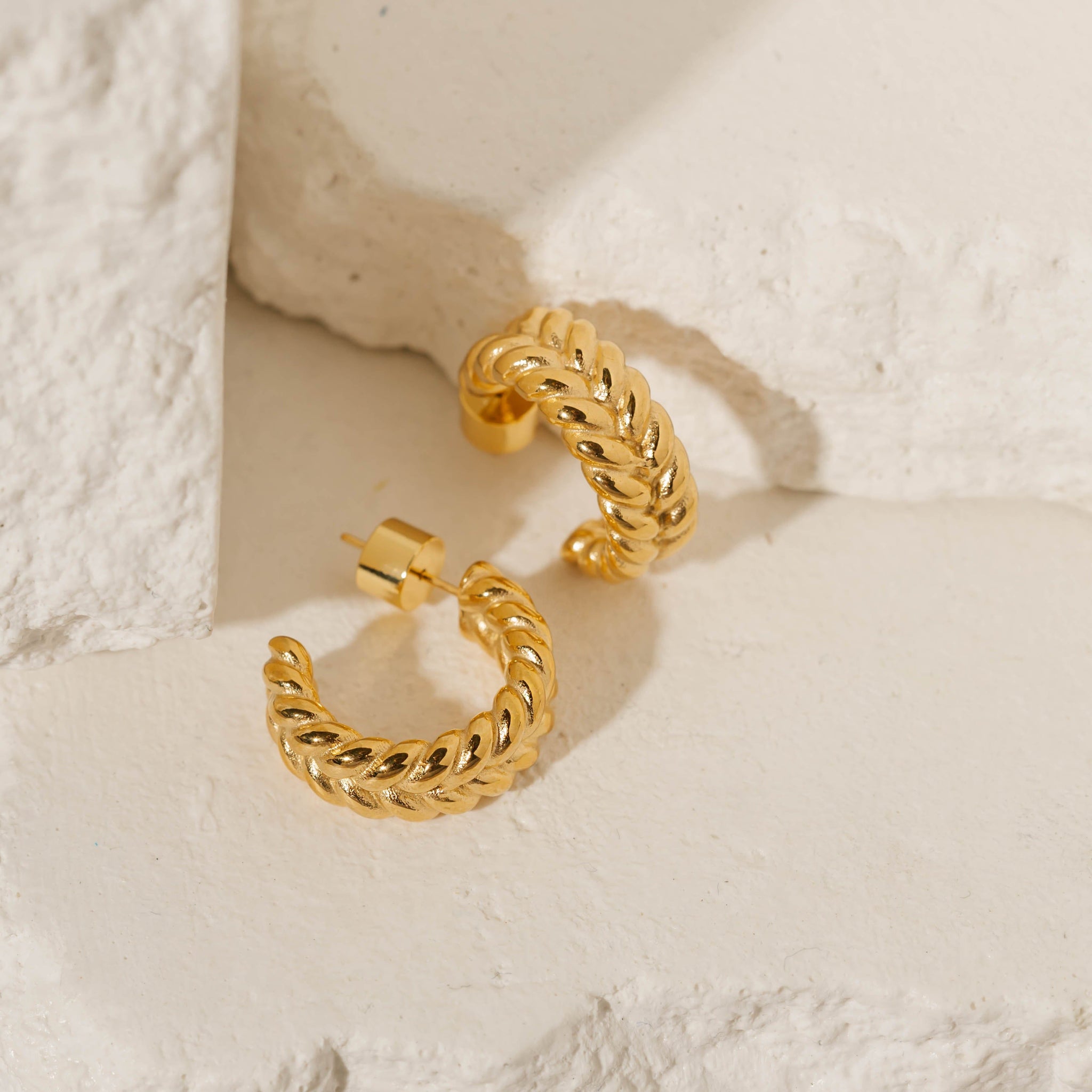 One Trenza Hoop earring lays on its side to show the cylindrical pushback closure and open hoop design as its pair stands tall to flaunt their braided design. 