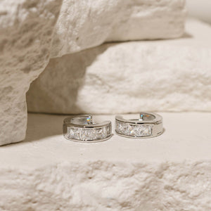 Platinum-layered Almeria Hoop earrings with cubic zirconia stones, elegantly displayed on a textured stone surface, showcasing a luxurious and refined style.