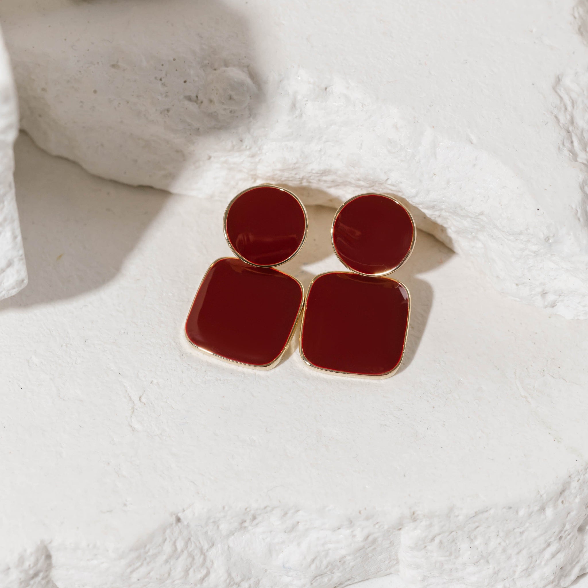 The Merlot Hoop earrings featuring geometric red enamel accents in a round and square design, framed in gold, elegantly displayed on a textured stone surface.