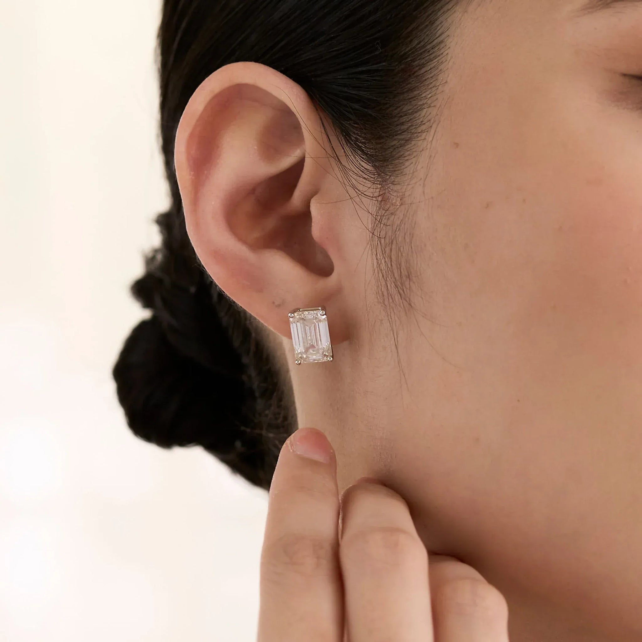 The Perfect Hoop Glass Stud earring worn on model with dark hair in low bun, showcasing a clear rectangular baguette-cut crystal on a silver stud, close-up side profile on soft cream background.
