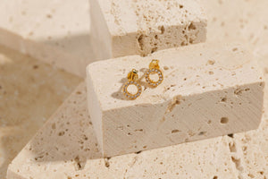 A pair of Infinito Stud earrings are displayed hanging side by side, the gold ring and crystal accents shimmering where the light reaches. 