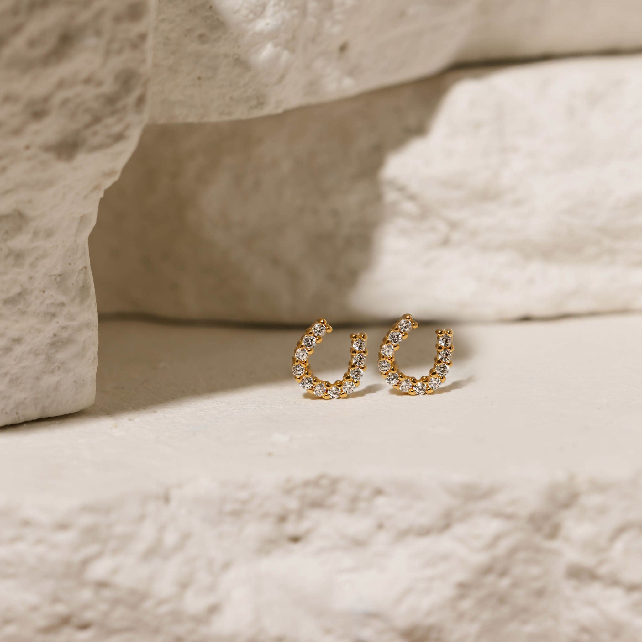  Close-up of The Lucky Studs, featuring a gold horseshoe design adorned with sparkling crystals, displayed on a textured neutral background.