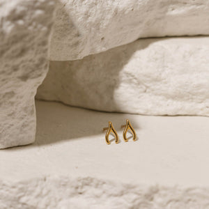 Close-up of The Make-a-Wish Studs, featuring a delicate golden wishbone design, displayed against a textured neutral background.