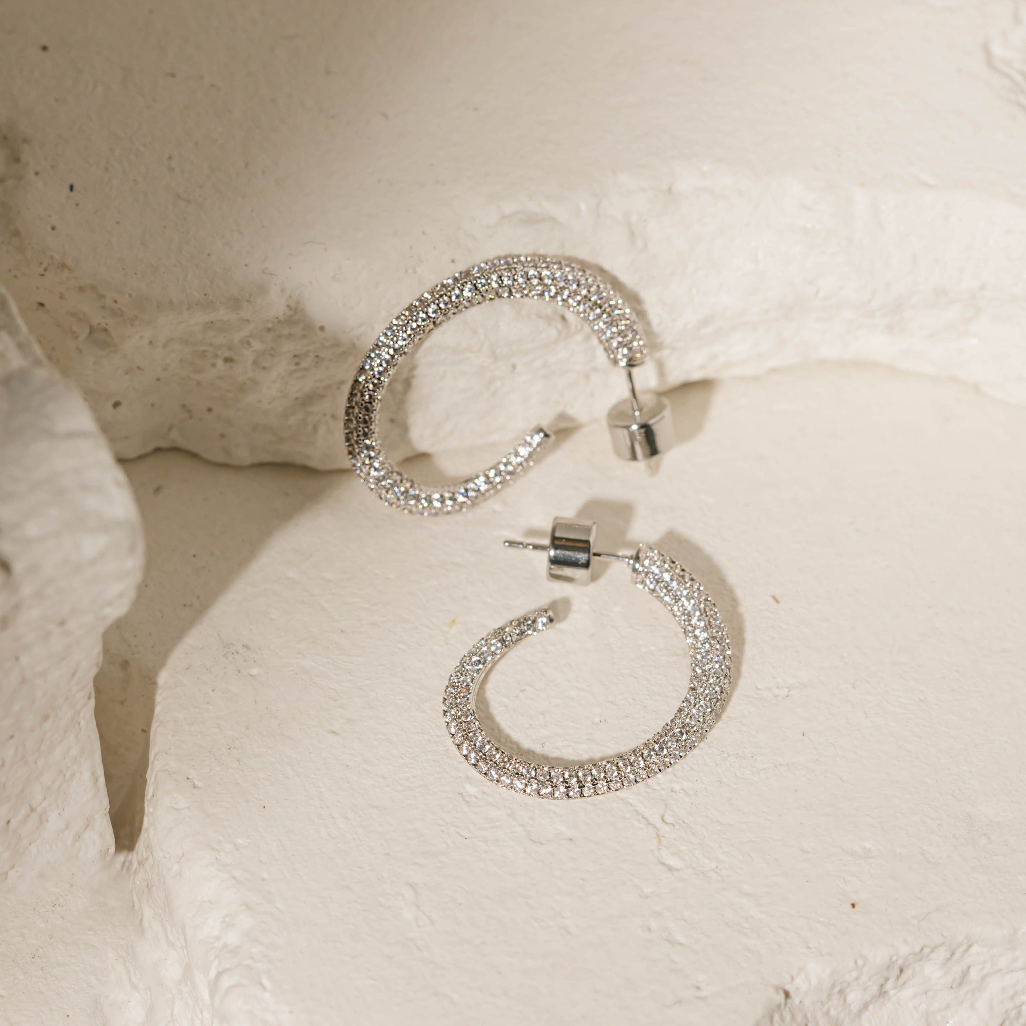 A pair of sterling silver Luxe Hoop earrings lies side-by-side against porous stone slabs to display the asymmetrical shape, sparkling crystal accents, and cylindrical push-back closures. 