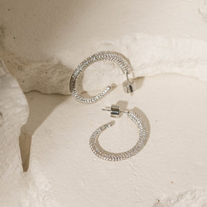 A pair of sterling silver Luxe Hoop earrings lies side-by-side against porous stone slabs to display the asymmetrical shape, sparkling crystal accents, and cylindrical push-back closures. 