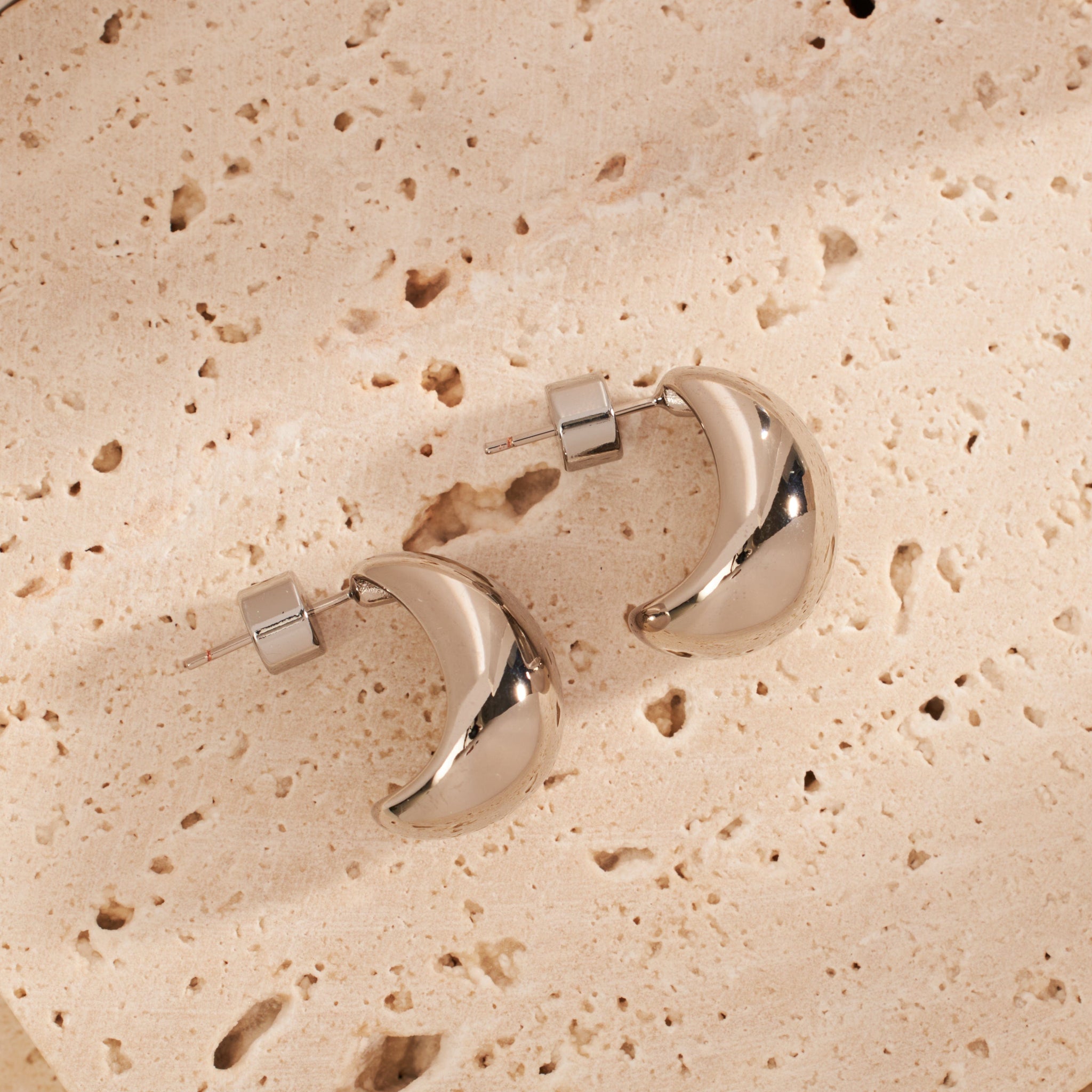 A pair of crescent-shaped Amadora Hoop Mini Platinum earrings is displayed laying side-by-side on a stone counter. 