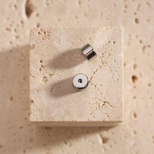 A pair of stainless steel Perfect Backings is displayed on a stone block, one sitting on the flat side to show the snug closure while the other lays on its side to show the cylindrical shape. 