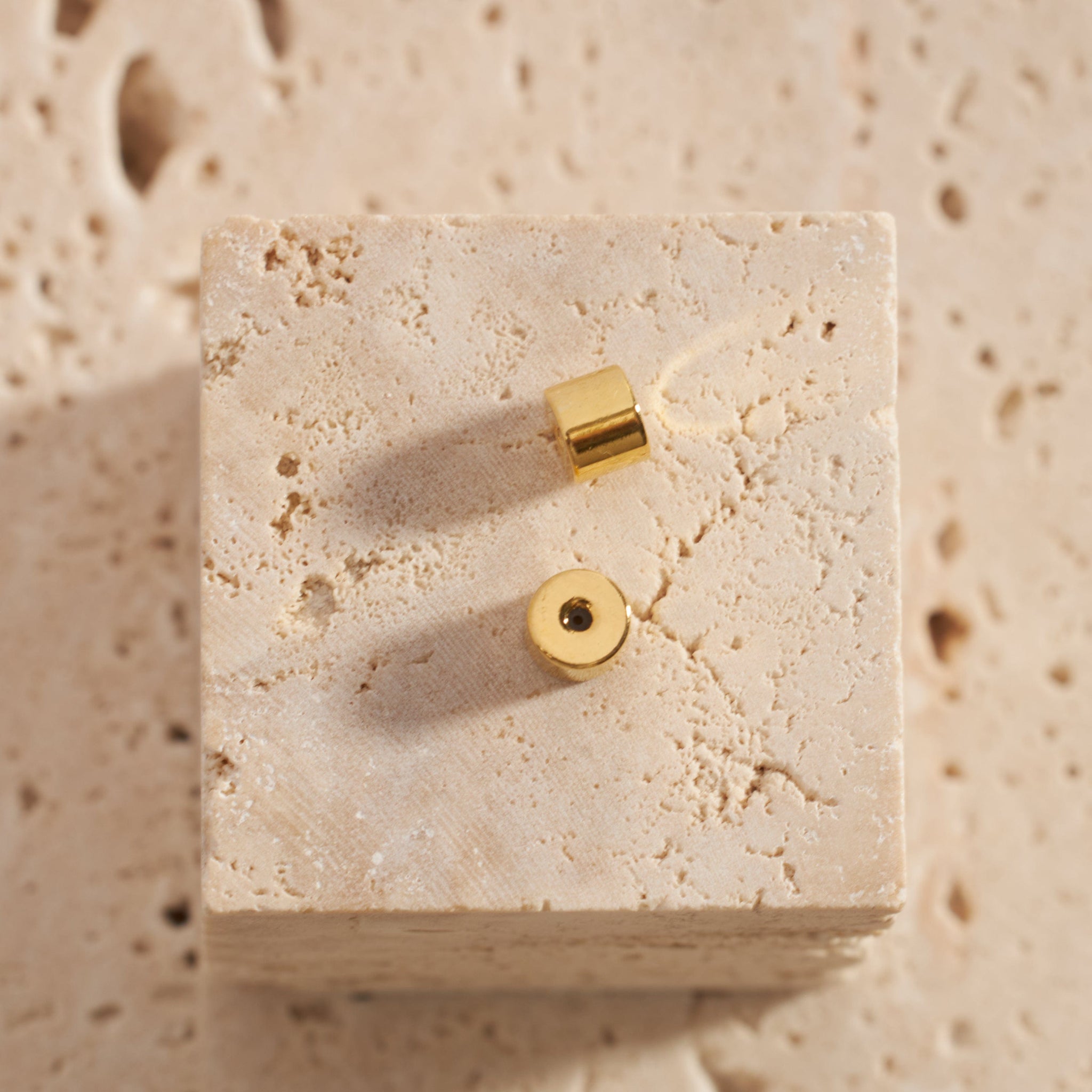 A pair of gold Perfect Backings is displayed on a stone block, one sitting on the flat side to show the snug closure while the other lays on its side to show the cylindrical shape. 