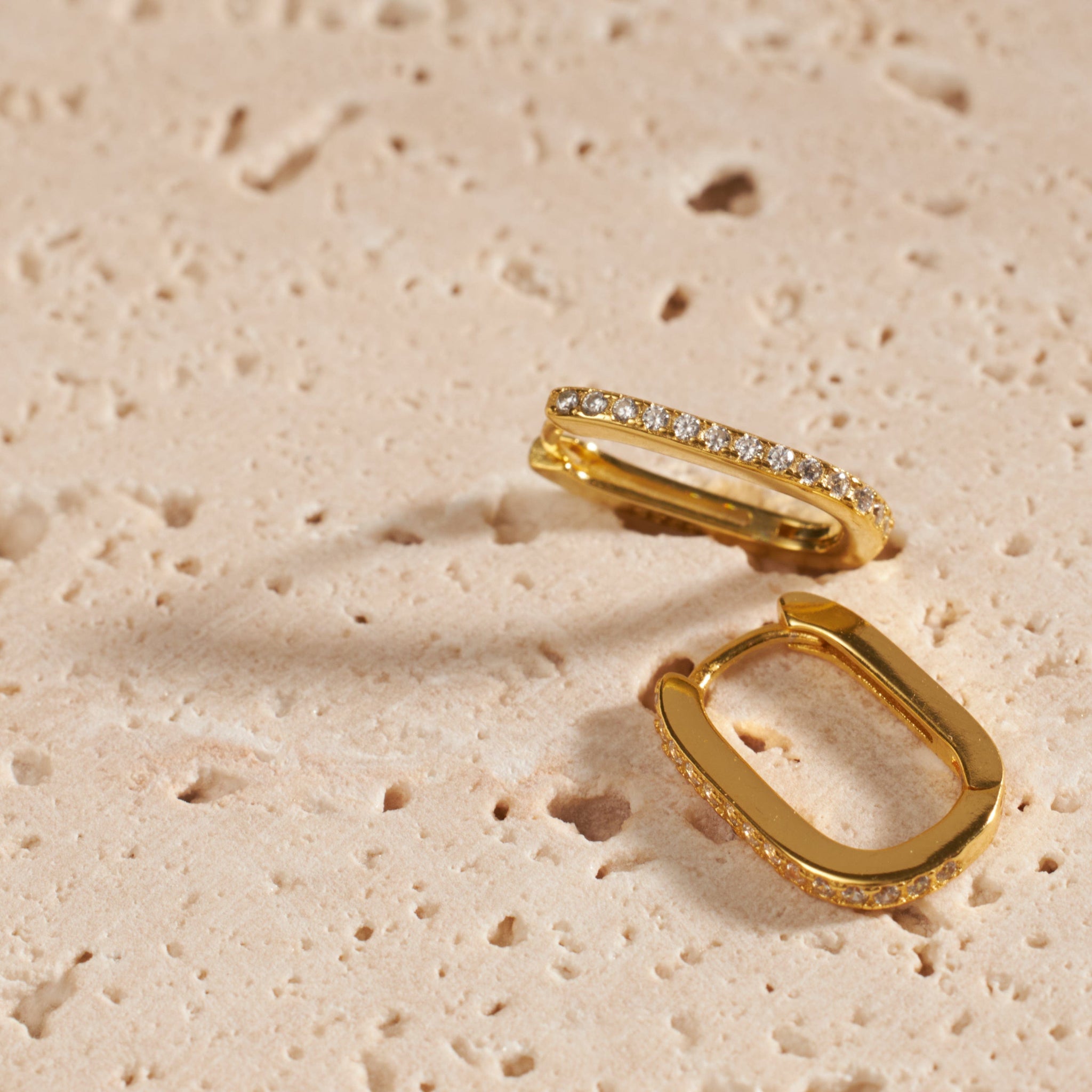 A pair of Positano Scintilla Huggie earrings is displayed side-by-side, one laying flat to show off the oval shape while the other sits on the side to display the sparkling crystal accents. 