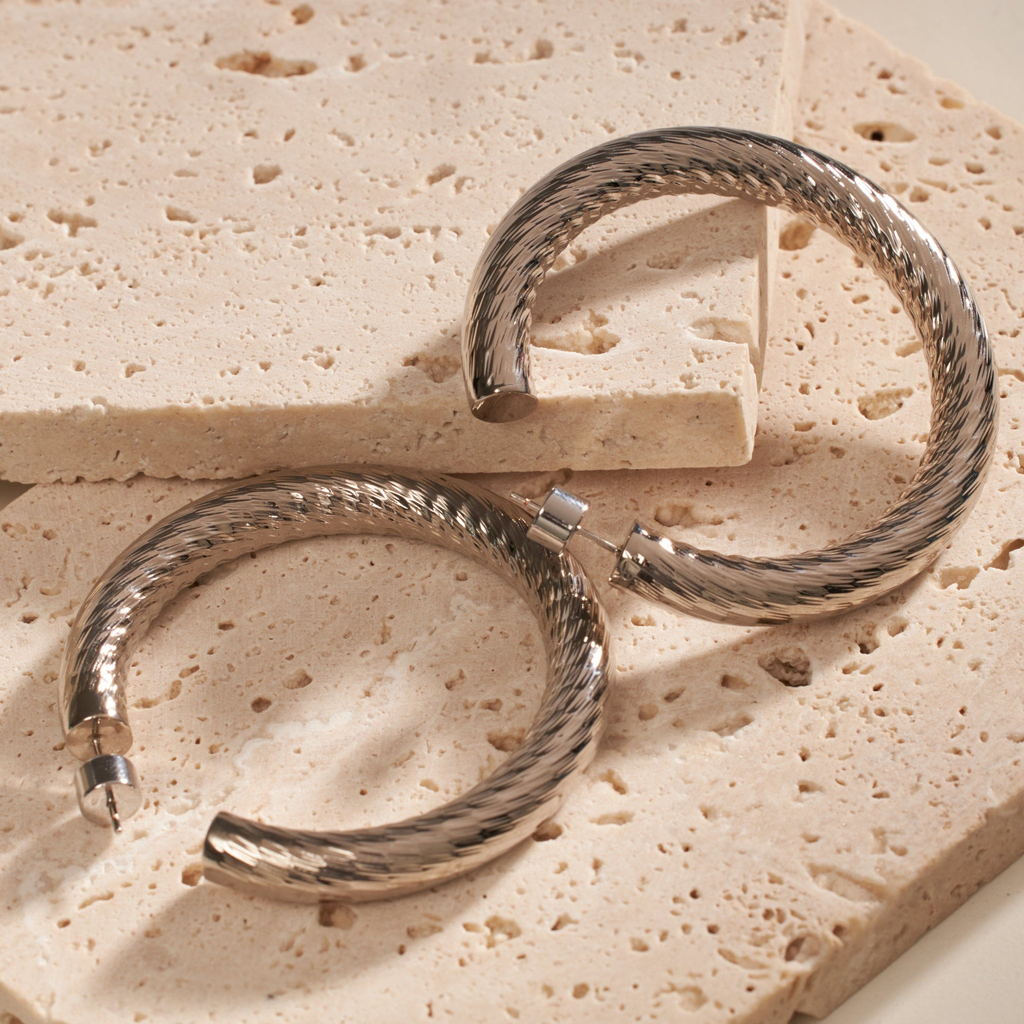  A pair of Madrid Hoop Platinum earrings lay side by side on two layered stone slabs, the close angle displaying the elegant rope-like texture, shining silver finish, and open hoop design. 