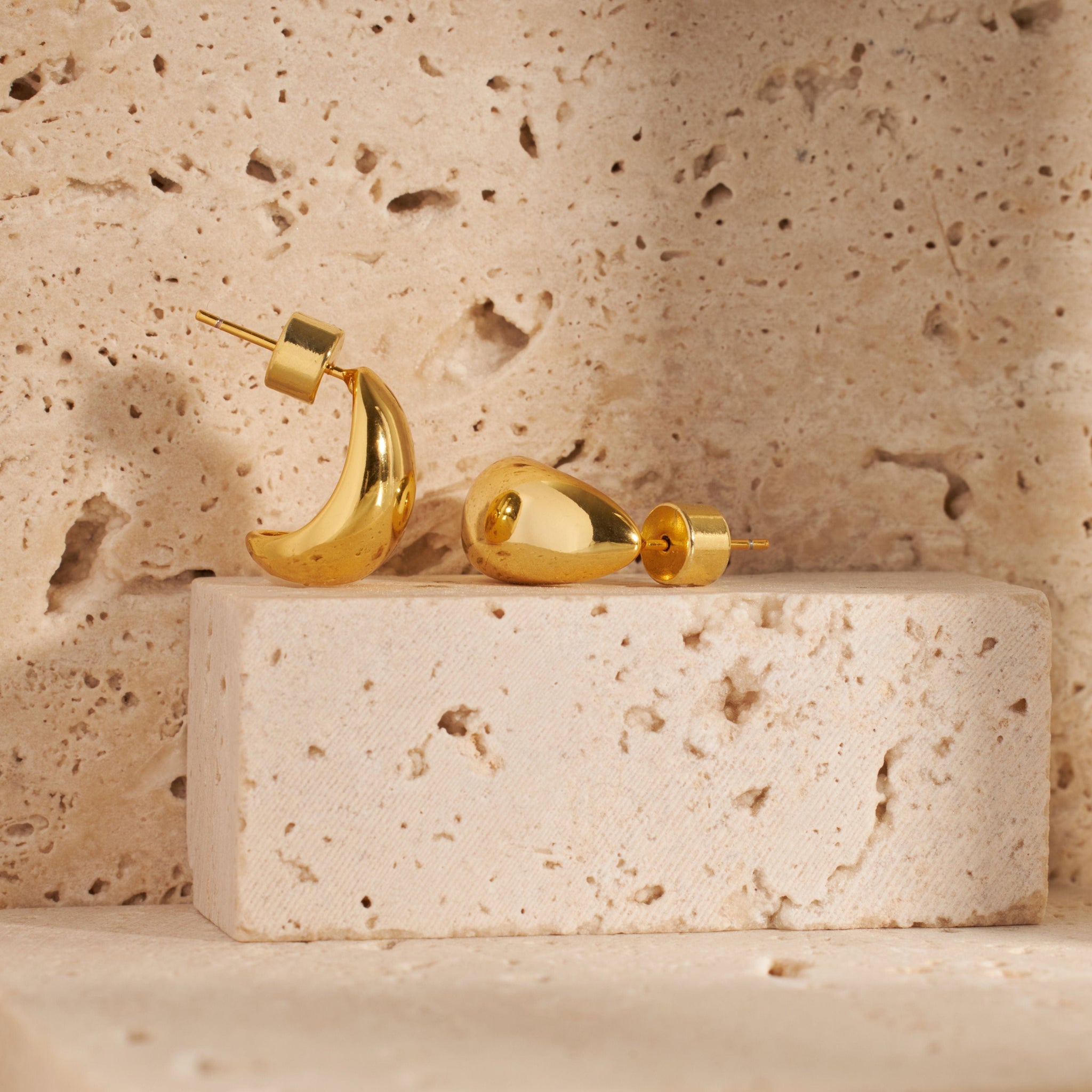 A pair of Amaldora Hoop Mini earrings lays on a raised, stone cube, the smooth golden surface gleaming in the light. 