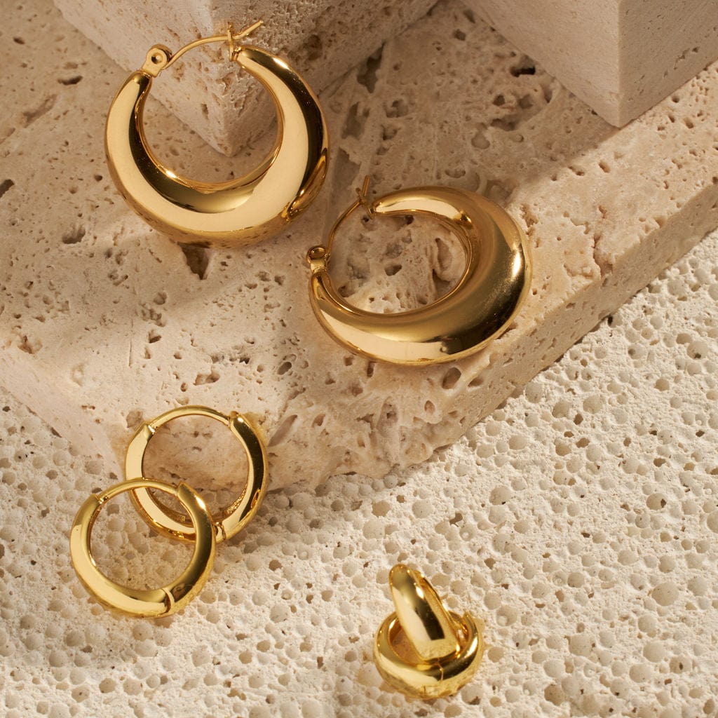 The Capri Hoop, Capri Huggie, and Capri Cuff are shown laying next to each other on tiered stone slabs, the sleek golden hoops of each earring gleaming brightly wherever the light hits them. 