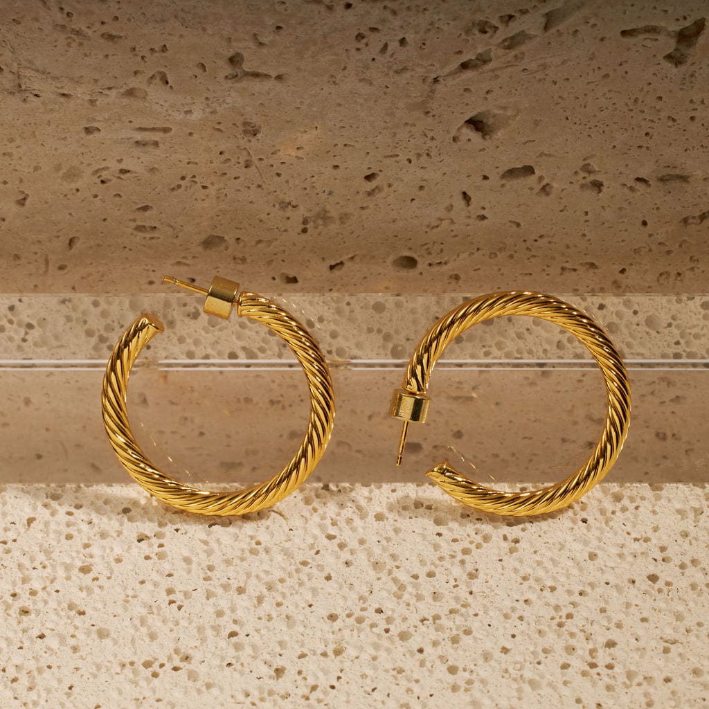 A pair of Bergamo Hoop earrings stand propped against a tiered stone surface to show off the spiraling ridged texture and open hoop design. 
