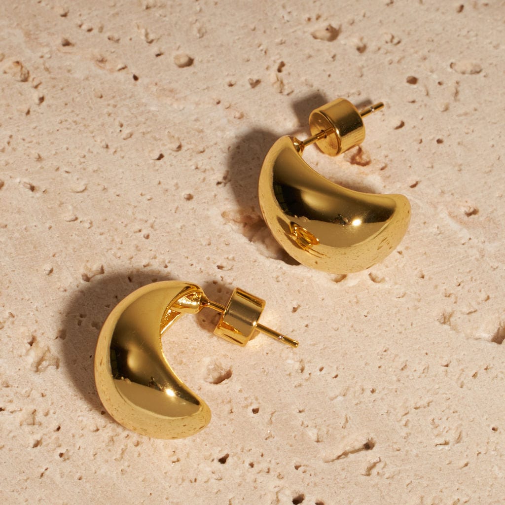 A pair of dome-shaped Amadora Hoop earrings lays side by side on a porous stone surface, the surroundings reflected in their smooth, golden curves.
