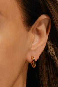 The Positano Hoop Mini worn on the ear, showing its compact, chunky oval shape in a glossy gold finish against brunette hair and warm skin tone.  By The Perfect Hoop.