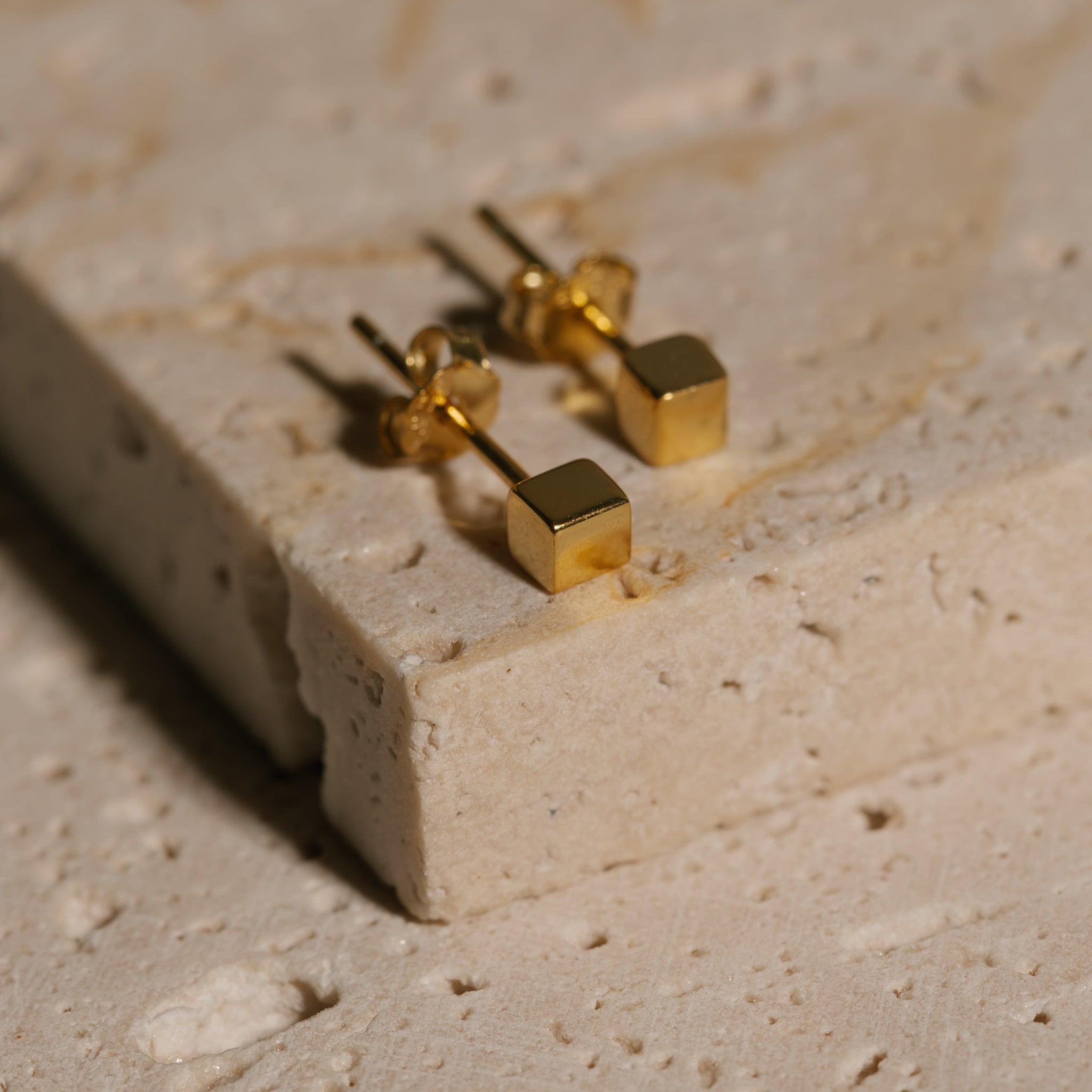A pair of golden, cube-shaped Cubo Stud earrings lay on a counter. 