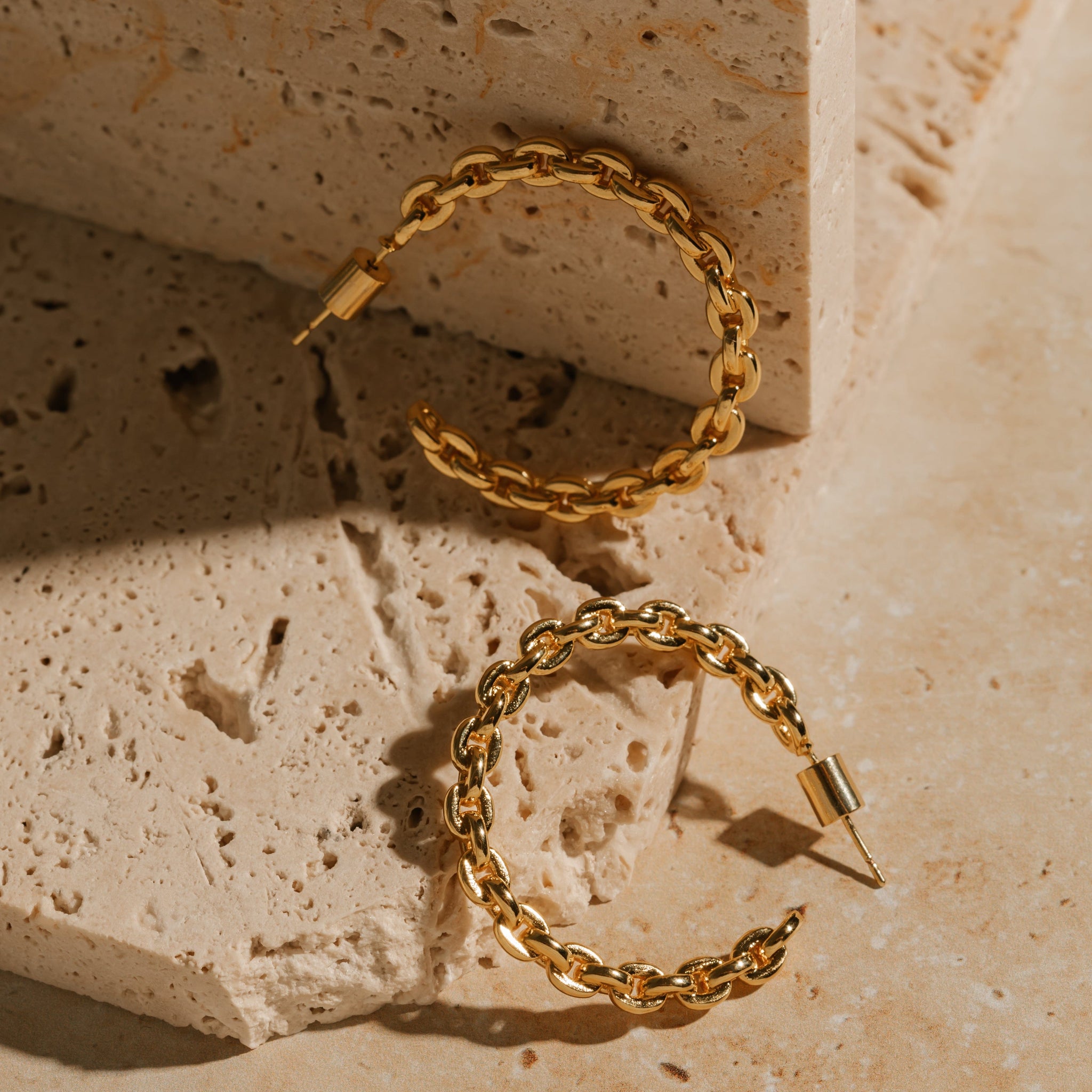 A pair of Catena Hoop earrings sits on a tiered stone slab, one earring propped up and glistening in the light while the other sits in the shadow of a raised block. 